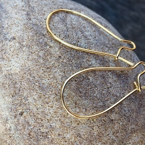 Op de afbeelding: Twee gouden oorhaken, gemaakt van metaal, liggen op een lichtbruine steen. De haken zijn ovaalvormig en hebben een kleine lus aan de bovenkant.