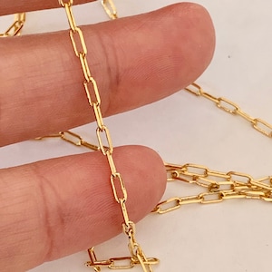 May include: A gold chain necklace with a delicate, rectangular link design. The chain is shown lying on a hand.