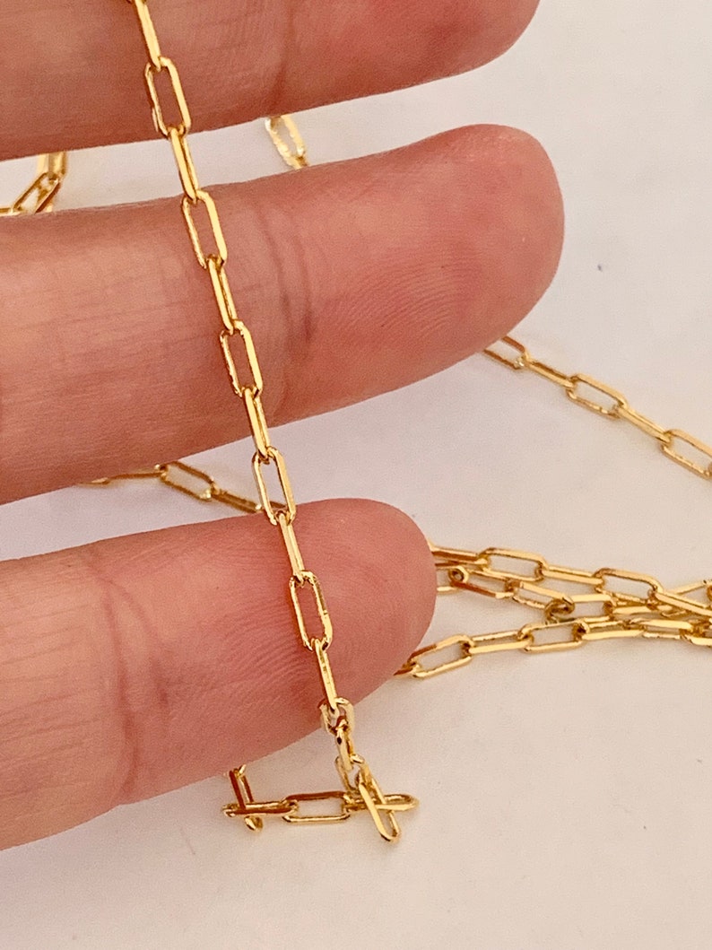 May include: A gold chain necklace with a delicate, rectangular link design. The chain is shown lying on a hand.