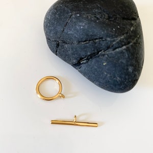 Puede incluir: Cierre de metal dorado con un anillo redondo y una barra recta. El cierre est&aacute; sobre una superficie blanca junto a una roca gris oscura.