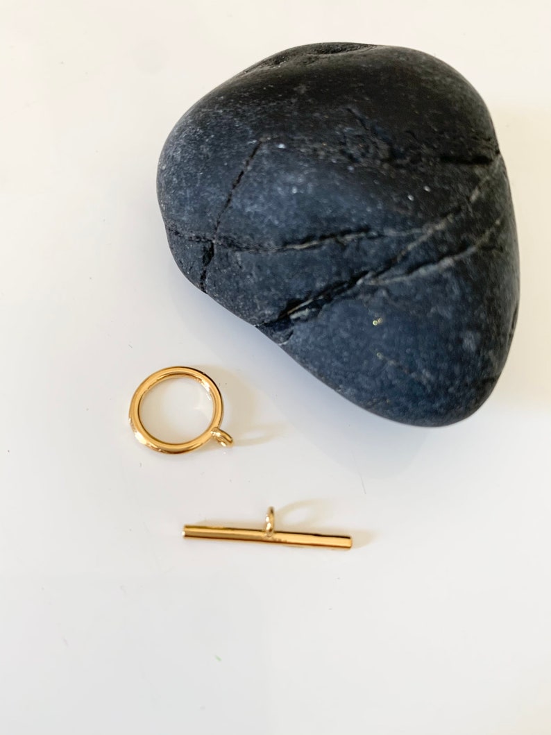 Puede incluir: Cierre de metal dorado con un anillo redondo y una barra recta. El cierre est&aacute; sobre una superficie blanca junto a una roca gris oscura.