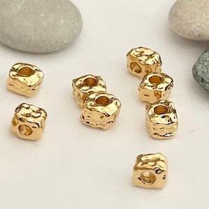 Könnte beinhalten: Goldfarbene Metallperlen mit einer strukturierten, unregelmäßigen Oberfläche. Jede Perle hat eine quadratische Form mit einem Loch in der Mitte.