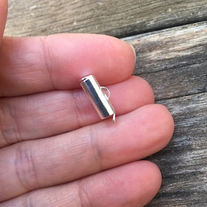 Op de afbeelding: Zilveren metalen schuifgesp met een lus aan het ene uiteinde. De gesp is cilindrisch van vorm.