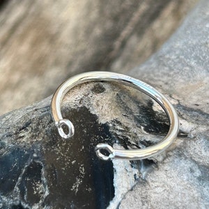 Könnte beinhalten: Ein silbernes Armreif-Armband mit zwei offenen Kreisen auf jeder Seite. Das Armband liegt auf einer grauen und schwarzen strukturierten Oberfläche.