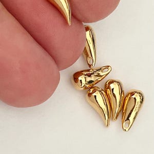 Pode incluir: Um grupo de seis pingentes em forma de lágrima de cor dourada com um pequeno laço no topo. Os pingentes estão dispostos em um cacho sobre uma superfície branca.