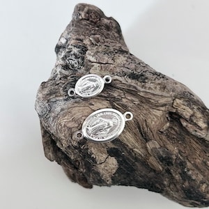 Può includere: Due ciondoli ovali in argento con l'immagine della Vergine Maria. I ciondoli hanno un anello su ogni lato per essere attaccati a una collana o un braccialetto.