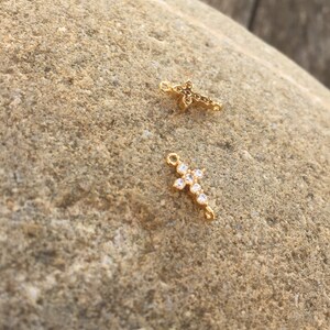May include: Two gold cross charms with clear crystal accents. The cross on the left is smaller and has a more intricate design. The cross on the right is larger and has a simpler design.