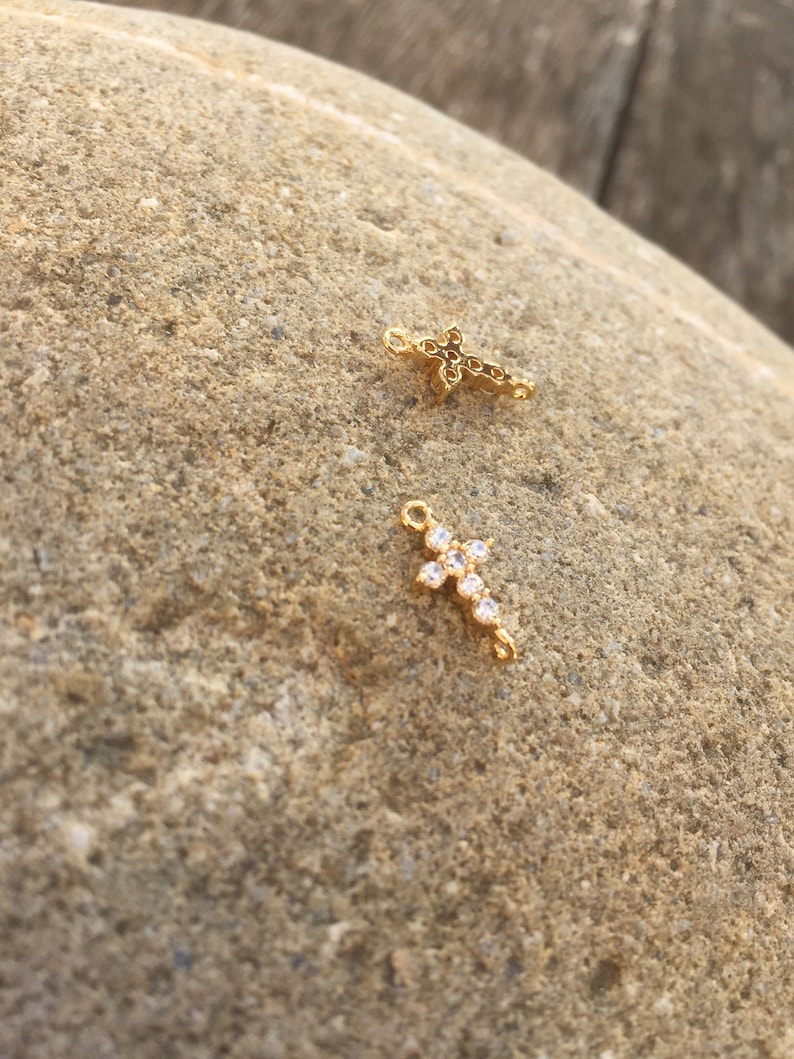 May include: Two gold cross charms with clear crystal accents. The cross on the left is smaller and has a more intricate design. The cross on the right is larger and has a simpler design.