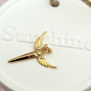 Peut inclure: Pendentif doré en forme de dague stylisée avec des ailes et des accents de cristal transparent. Le pendentif est sur une surface blanche avec le mot "Sunshine" imprimé dessus.