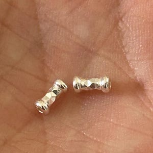 May include: Two small, silver-colored, dumbbell-shaped beads with faceted surfaces. The beads are displayed against a blurred background, suggesting they are jewelry components or craft supplies.