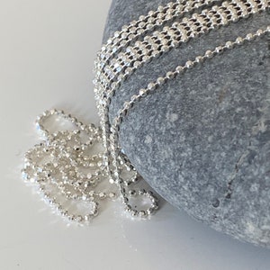Peut inclure: Un collier en chaîne argentée avec un design délicat de chaîne à billes. La chaîne est posée sur une pierre grise.