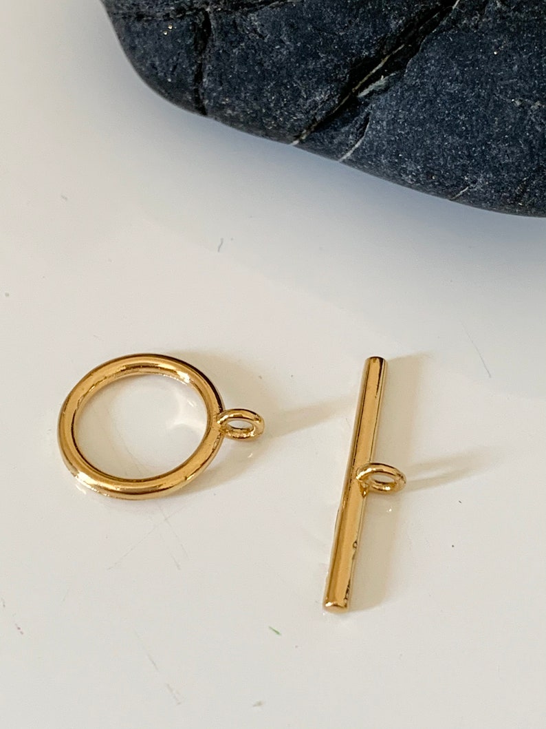 Puede incluir: Cierre de palanca de metal dorado con un anillo redondo y una barra recta. El cierre se utiliza para la fabricaci&oacute;n de joyas.