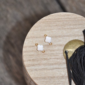 Puede incluir: Dos conectores de metal dorado con dise&ntilde;os de tr&eacute;bol de cuatro hojas de esmalte blanco. Los conectores est&aacute;n sobre una superficie de madera.