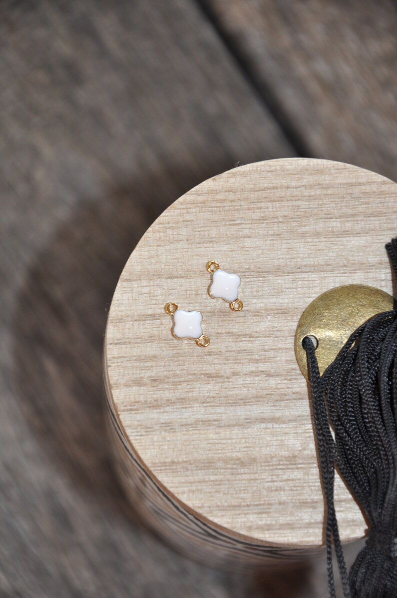 Puede incluir: Dos conectores de metal dorado con dise&ntilde;os de tr&eacute;bol de cuatro hojas de esmalte blanco. Los conectores est&aacute;n sobre una superficie de madera.