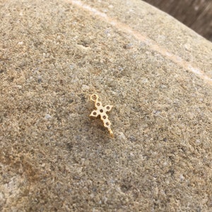 May include: A small gold cross charm with a clear stone design. The charm is on a rough, brown rock surface.