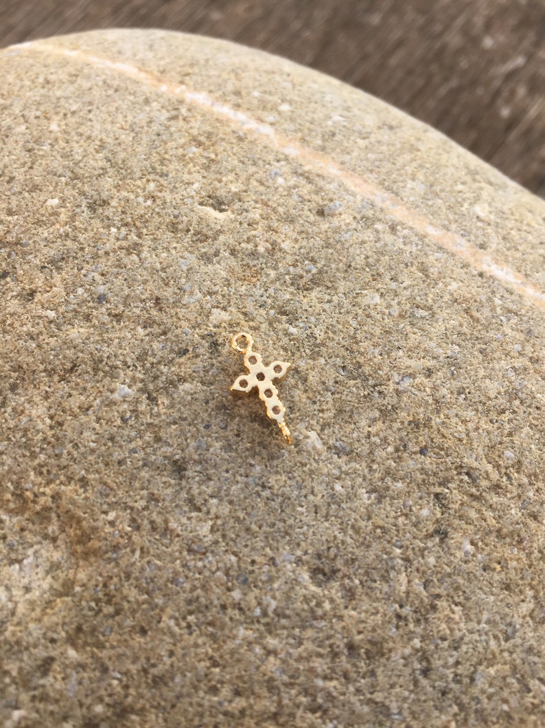 May include: A small gold cross charm with a clear stone design. The charm is on a rough, brown rock surface.
