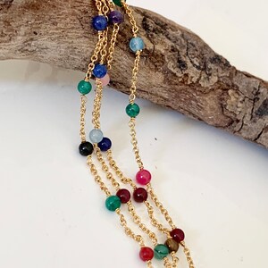 Op de afbeelding: Een gouden ketting met gekleurde edelsteen kralen. De kralen zijn in verschillende kleuren, waaronder rood, groen, blauw en geel.