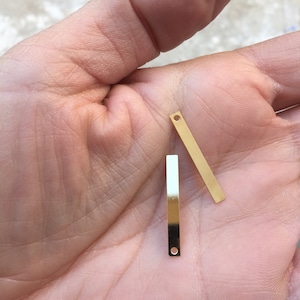 Pode incluir: Duas formas de barra de metal de cor dourada com um pequeno furo em cada extremidade. As barras medem aproximadamente 2,5 cm de comprimento.