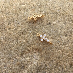 May include: Two gold cross charms with clear crystal accents. The charms are on a rough, brown stone surface.