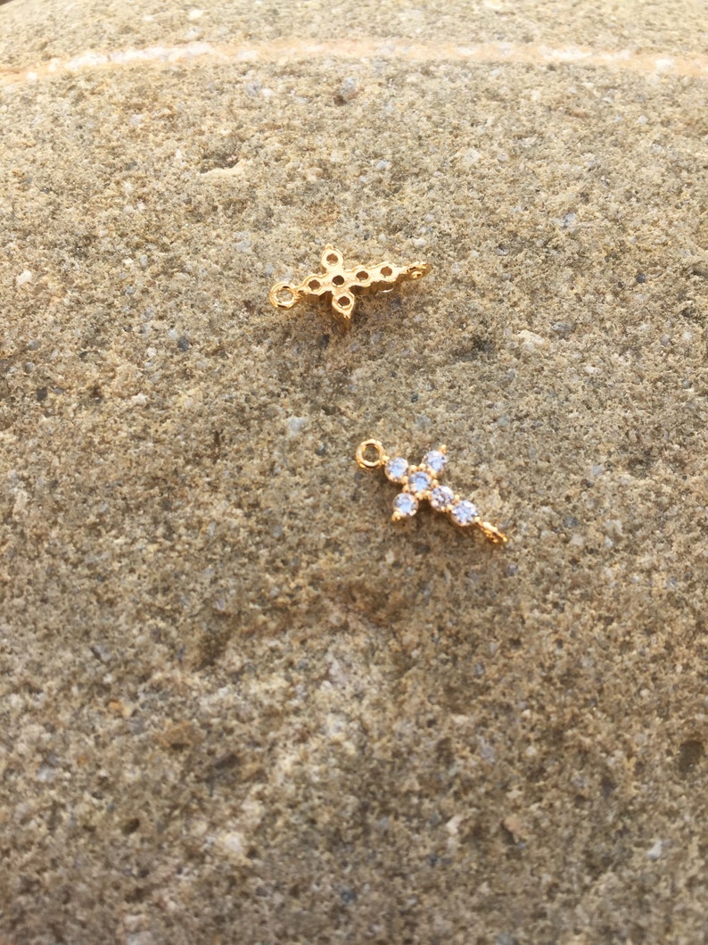 May include: Two gold cross charms with clear crystal accents. The charms are on a rough, brown stone surface.