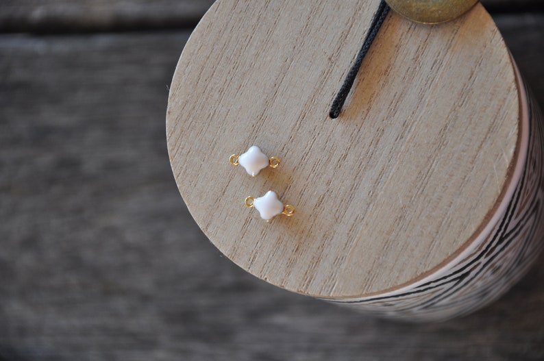 Puede incluir: Dos encantos de tr&eacute;bol de cuatro hojas blancos con anillos de salto dorados. Los encantos est&aacute;n sobre una superficie de madera.