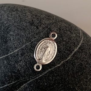 Peut inclure: Un pendentif ovale argenté avec une image religieuse et du texte. Le pendentif comporte deux petites boucles pour l'attache. L'image représente une figure aux bras tendus. Le pendentif est sur une surface sombre et texturée.