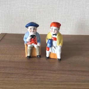 May include: Two ceramic figurines of people sitting in chairs. The person on the left is wearing a blue hat, blue jacket, red waistcoat, and brown trousers. The person on the right is wearing a red hat, yellow jacket, white apron, and brown trousers.