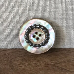 May include: A vintage oval-shaped mother-of-pearl button with a black bead border and four holes in the center.