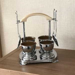 May include: A vintage chrome egg cup holder with four cups and two spoons. The holder has a cream-coloured handle and is set on a silver-coloured base. The egg cups are shiny and metallic, ideal for serving eggs at breakfast or brunch.