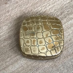 May include: A small, rectangular, gold-colored compact with a textured, cobblestone-like pattern on the lid. The compact is closed and sits on a wooden surface. The overall design suggests a vintage or antique style.