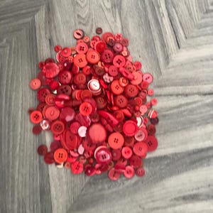 May include: A collection of red buttons in various shades and sizes. The buttons are scattered, showcasing a range of shapes, including round, square, and those with multiple holes. The color palette includes shades from light pink to deep crimson.