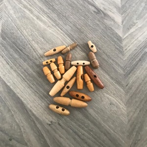 May include: A collection of wooden toggle buttons in various shades of brown. The buttons have two holes and are in different sizes and shapes.