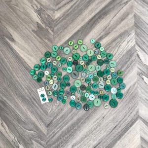 May include: A collection of green buttons in various shades, including teal, emerald, and olive, scattered on a wooden surface. The buttons vary in size and shape, with two- and four-hole designs. A small card with two buttons is also present.