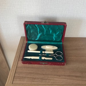 May include: A red velvet box with a green satin lining, containing a set of vintage grooming tools. The tools include a small brush, a pair of scissors, a nail file, and a small jar.
