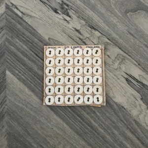 May include: A grid of off-white, round buttons with two holes, presented on a vintage cardboard backing. The text "MADE IN 22 LINES, ENGLAND" is visible. The buttons are set against a grey, wood-grain background.
