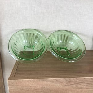 May include: Two vintage green glass bowls with a ribbed design. The bowls are round with a decorative base and are displayed on a wooden surface. The glass has a slight glow, suggesting they are made of uranium glass.