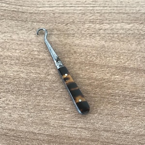 May include: An antique metal hook tool with a curved end and a tortoiseshell patterned handle. The tool is set against a wooden background, highlighting its vintage design and craftsmanship.