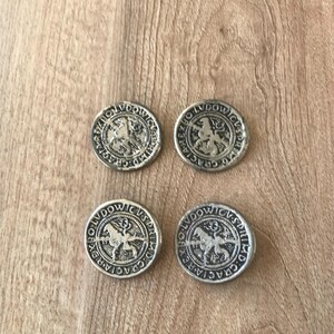 May include: Four silver-colored coins with a raised design of a horse and rider, surrounded by text. The coins are arranged in a square on a wooden surface. The coins appear to be antique or vintage.