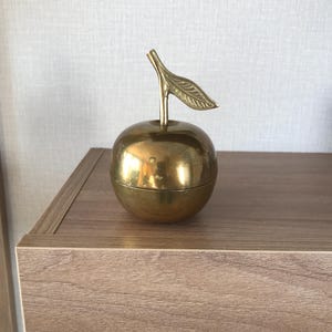 May include: A brass apple-shaped container with a stem and leaf detail. The container is round and has a shiny, reflective surface. The leaf and stem are attached to the top of the apple. The container is sitting on a wooden surface.
