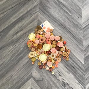May include: A collection of assorted buttons in various sizes and colors, including shades of pink, brown, and yellow. Some buttons have two or four holes, while others have a shank. A tag with text is visible among the buttons.