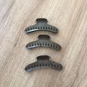 May include: Three black metal hair clips with a curved shape and a row of teeth along the inside edge. The clips are arranged in a vertical stack.