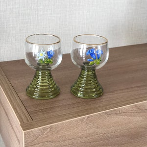 May include: Two clear glass wine glasses with green stems and bases. Each glass has a gold rim and a painted floral design featuring blue and white flowers.