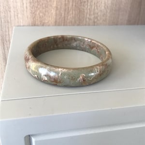 May include: A round, jade-colored bracelet with a mottled pattern of brown and white. The bracelet is smooth and polished, resting on a white surface. This piece of jewelry is a classic accessory.