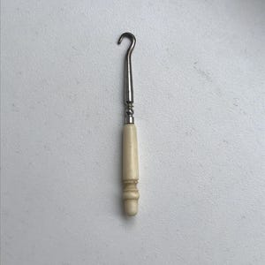 May include: A vintage crochet hook with a curved metal hook and a cream-colored handle. The handle has a decorative, ridged design. The hook is a tool used for creating crocheted items, such as blankets, scarves, and garments.