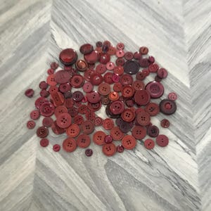 May include: A collection of various red buttons in different sizes and shades, from deep burgundy to bright cherry. The buttons are scattered on a light gray and white chevron patterned surface. The buttons have two or four holes.