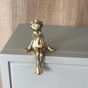 May include: A gold-coloured frog figurine with outstretched arms and legs, sitting on a grey surface. The frog has detailed features and a smiling expression. The figurine is likely a decorative item.