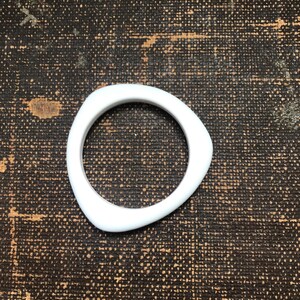 May include: A white, triangular-shaped bangle bracelet. The bracelet has a smooth, rounded design and a hollow center. The background is a dark, textured surface.