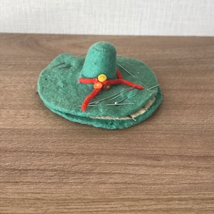 Peut inclure: Un coussin à épingles en feutre vert en forme de sombrero avec un ruban rouge et des boutons jaunes, orange et rouges. Le coussin à épingles a plusieurs épingles qui en sortent.
