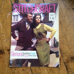 以下が含まれることがあります： 1963年10月号のヴィンテージのStitchcraft雑誌の表紙で、ニットを着たカップルが写っています。雑誌のタイトルはピンクの文字で、表紙のテキストは「Glenora Sparkling Tweed for Autumn Fashion」です。