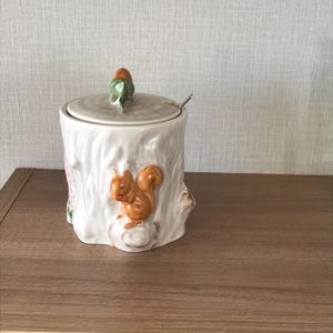 May include: A white ceramic sugar bowl with a lid and a small spoon. The bowl is shaped like a tree stump with a detailed orange squirrel on the front. The lid has a green leaf and orange acorn handle. A charming addition to any kitchen.